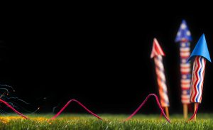Three red, white, and blue fireworks stuck in the ground and connected to a lit fuse.