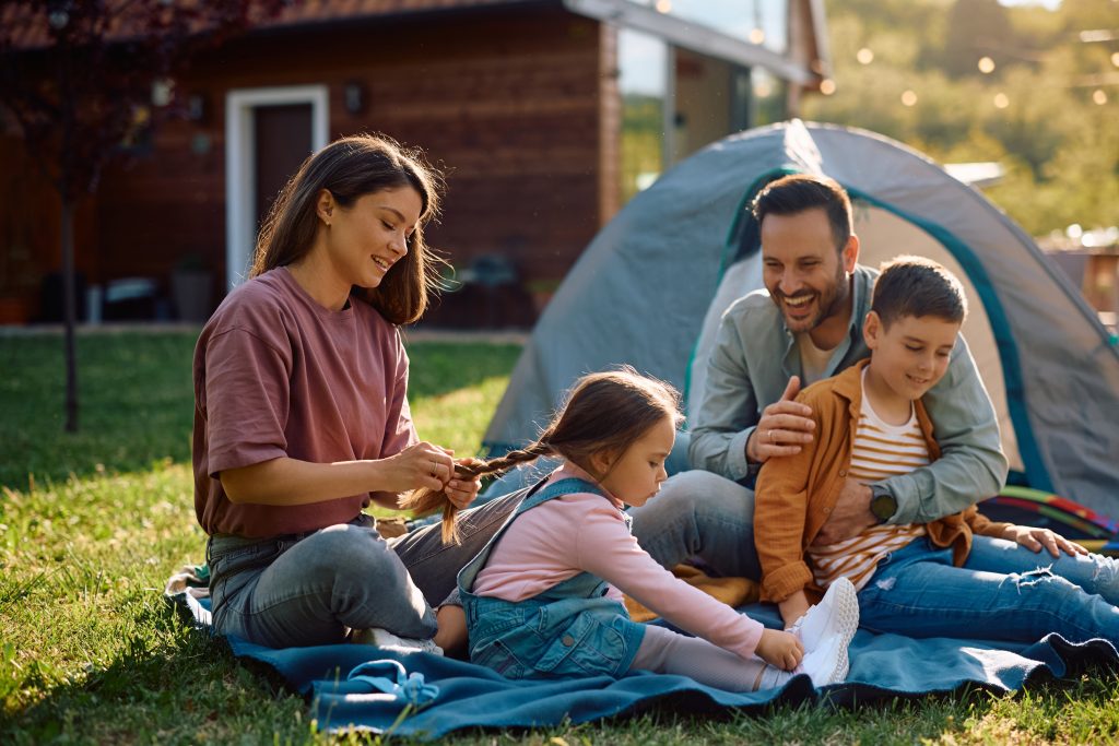 Parents and two children sitting in a yard in front of a tent, the mother braiding the daughter's hair.