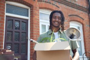 A man stands, smiling, in front of an apartment building with moving box in his hands.