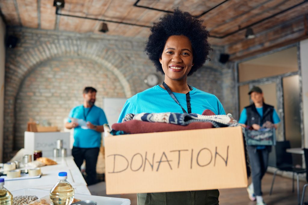 A volunteers holds a box  labeled "Donation" while smiling.