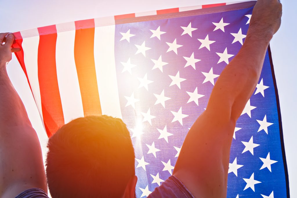 A person holding up an American flag in front of the sun.