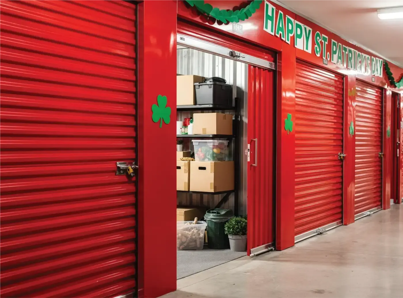 A long hallway in a modern indoor self-storage facility with bright red roll-up doors. One unit is open, revealing neatly stacked cardboard boxes and storage bins on metal shelving. The facility is decorated for St. Patrick's Day with green shamrock decals on the door frames and festive green garlands and a "Happy St. Patrick's Day" banner hanging across the top of the units.