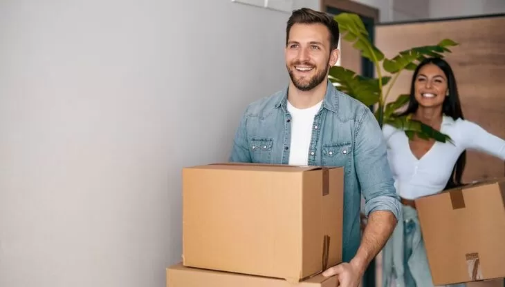 A couple is in the process of moving, with the man in the foreground carrying boxes and the woman in the background, both appearing happy and active.